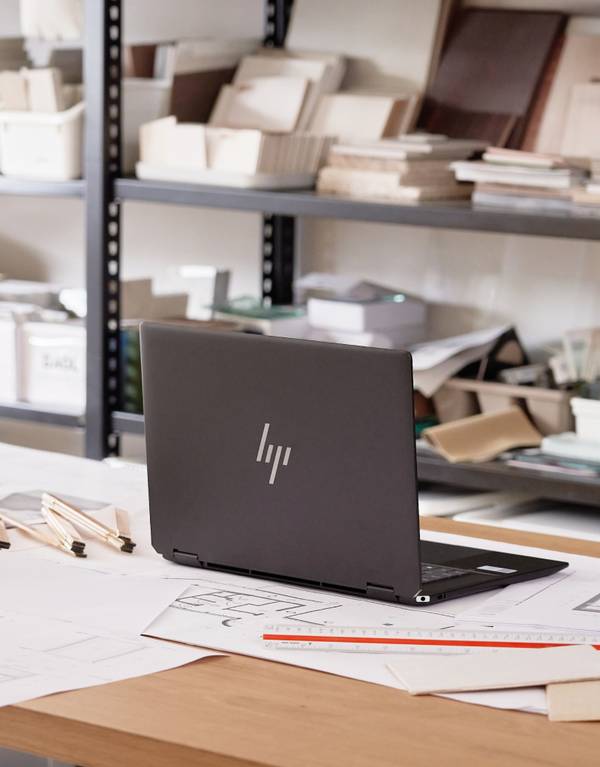 HP 스펙터 x360 2-in-1 노트북이 설계 도면이 펼쳐진 책상 위에 놓여 있고, 뒤편 선반에 작업 자료들이 정리된 모습.